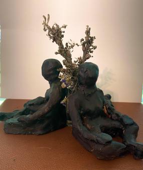Three black clay figural forms sitting with their hands in their laps against a small glass vase filled with dried flowers. This is positioned on a brown leather cloth. 