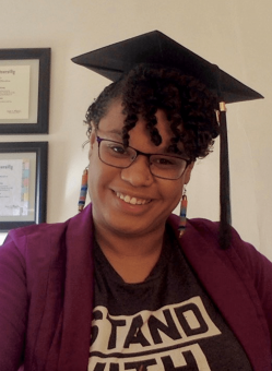 Christine smiles while wearing a graduate cap. Christine wears glasses and a shirt that reads "Stand."