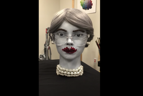 Video still of foam head painted grey with grey eyes, long eyelashes, and glasses. The bust has straight grey hair pulled back, is wearing bright red crystal beads for lipstick, and has on a chunky pearl necklace and earrings. The face has brown eyebrows made from coils. 