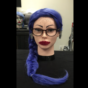 Head bust with pale skin and long blue hair styled in a braid. The face is wearing glasses, has black drawn on eyebrows, black/brown glasses, and has red crystals as lipstick. Drawn coming down from the forehead is a lotus like mandala flower design. They hasve grey fringy earrings. 