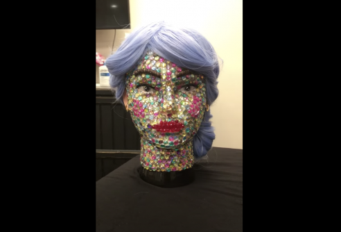 Face bust with light grey eyes, long eyelashes, and light blue/grey hair pulled back. The face is covered in multicolored crystal gems.  The lips are covered in bright red crystal beads. 