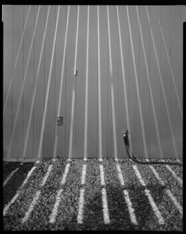 Black and white photo of pebbles in front of a fence. Long skinny rays of sunlight shine down the fence and onto the pebbles.