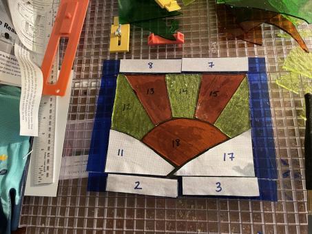 aturing an orange sun positioned between two green hills. Orange and yellow sunbeams radiate out of the sun. Royal blue stained glass creates a rectangular border. In this progress photo, the glass is on a cutting mat with various tools. White numbered cut outs of paper are positioned on the glass so the artist can cut and piece the glass.