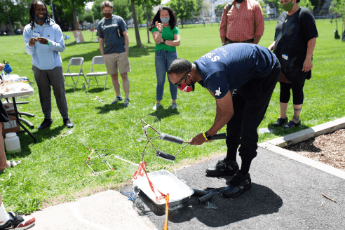 Surrounded by a small group of people in a grassy park, a man bends down working on a makeshift contraption. 
