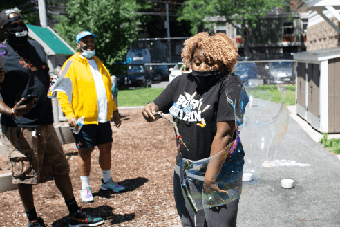 A woman looks surprised while making a huge bubble using a bubble wand made from a stick.