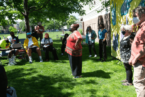 Outside a woman with her hands on her hips stands in front of a group of people sitting in metal folding chairs filling out paperwork.