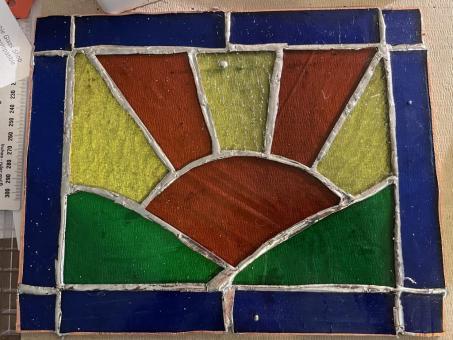 Photograph of a stained glass rectangle featuring an orange sun positioned between two green hills. Orange and yellow sunbeams radiate out of the sun. Royal blue stained glass creates a rectangular border. 