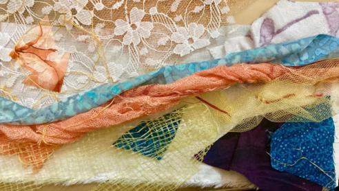 Mixed Media. Several pieces of fabric overlay each other on a yellow background. Assorted colors of fabric are twisted and create a horizontal composition. The colors include blue, yellow orange, brown, and white. Underneath three quarters of the fabric, starting at the bottom is a white piece of fabric with a frayed edge. The fabrics vary in texture and print including lace and mesh. 