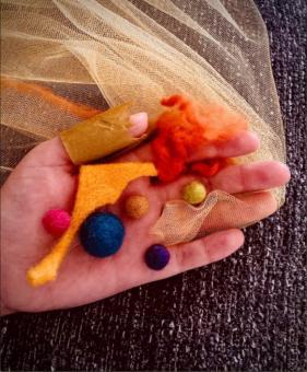 In the center of the piece is a hand facing palm up. The hand is holding several differently sized felt balls (blue, pink , brown, purple and yellow). In between the pink and the blue balls is a long shape cut out of light orange felt. Wrapped around the pointer finger of the hand is bright orange yarn. Wrapped around the thumb is a cutout piece of brown paper. Wrapped over the ring finger is a yellow piece of mesh fabric which extends upward beneath the other fingers and out past the edge of the frame. 