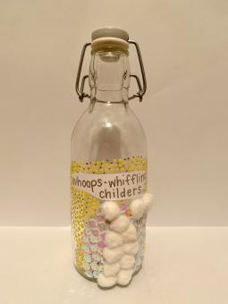 Mixed media. A glass bottle with a built-in stopper. On the bottle is a piece of paper with the words “gloriumptious human beans” written in silver. Surrounding these words are pink and white sequins and white pompoms and on the outside of those are navy blue and silver. In the bottle can be seen small slips of paper that appear to have words written on them.