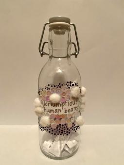 Mixed media. A glass bottle with a built-in stoppers. The bottle has a piece of paper with the words “whoops-whiffling childers” written in silver. Surrounding the words are yellow and silver dots which also extend down to surround white pompoms and pink and white sequins which are below the words. 