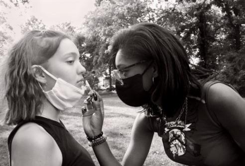 A black and white photo taken by Sofia Dushku, a student in the Lesley Pre-College program. It shows a masked Black woman putting lipstick on a white woman, who has her mask below her mouth.