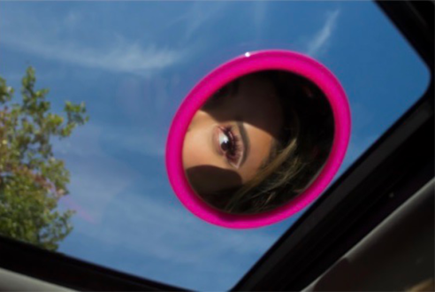 A photo by Katie Medrano Escobar. It shows a girl looking at her eye in a small mirror attached to a car window.