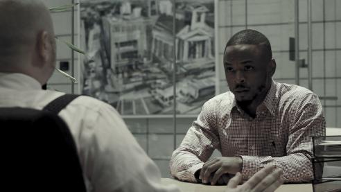 A young man in dress clothes sits at a desk looking at another person