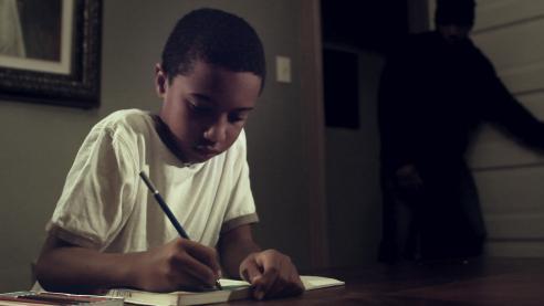 A young child sits at a table writing on paper while the shadow of a person is seen across the room