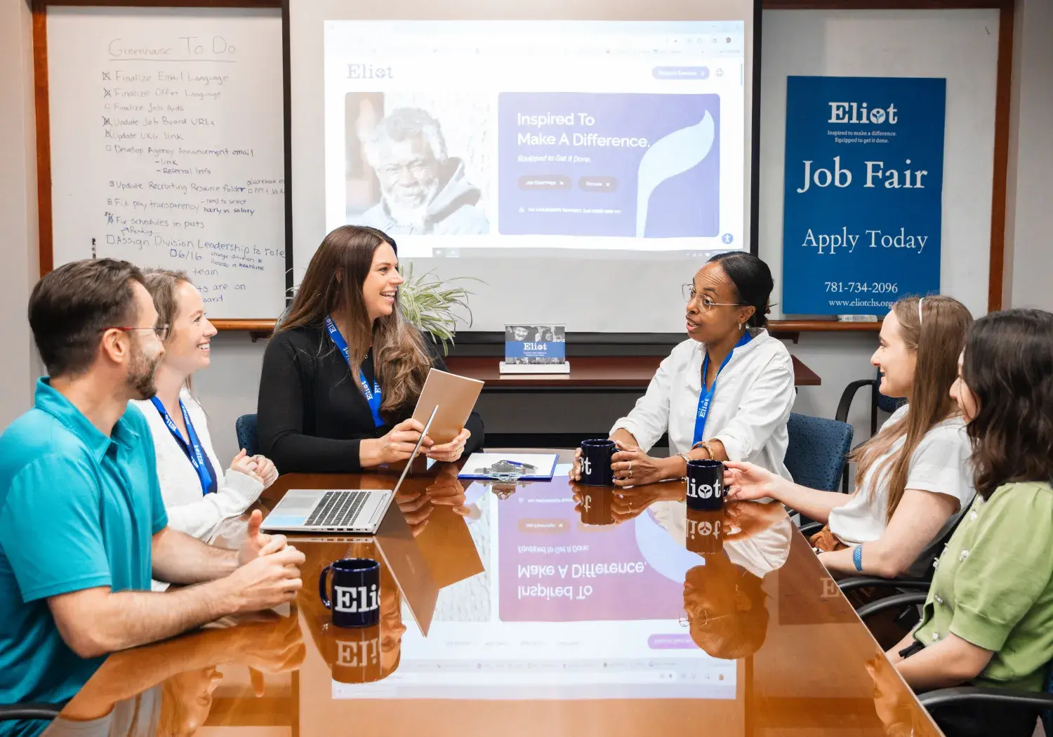 A diverse group of six people are seated around a polished wooden conference table, engaged in a discussion. A laptop, more "Eliot" mugs, and papers are on the table. Behind them, a large screen displays a website or presentation with the word "Eliot" and "Inspired To Make A Difference." To the left of the screen, a whiteboard lists "Crossover To-Do" items. On the far right, a blue poster advertises an "Eliot Job Fair."