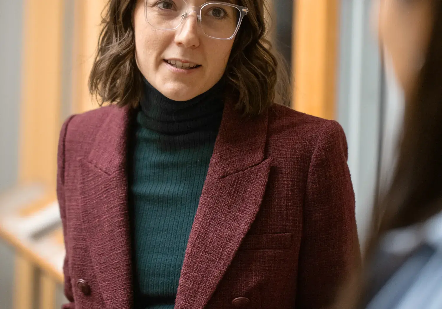 A woman with short, wavy brown hair and clear-framed glasses wears a dark red blazer over a dark green turtleneck and a plaid skirt. She is looking directly at the camera with a slight expression of speaking, her mouth slightly open and her right hand open as if gesturing. The shoulder and arm of another person are visible in the blurred foreground on the right. In the background, there are vertical wooden panels.