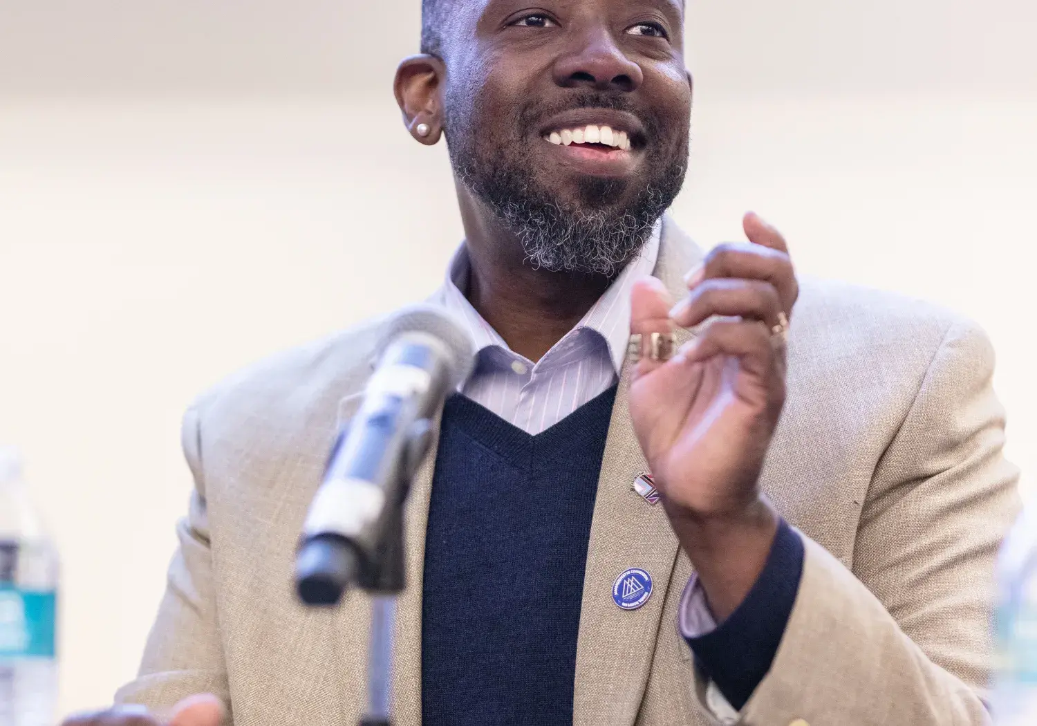 A professionally dressed man smiles warmly while speaking at a panel discussion. He wears a beige blazer over a navy sweater and collared shirt, with two pins on his lapel. He gestures with his hands as he speaks into a microphone on a stand. A water bottle sits on the table in front of him. The setting appears to be an indoor conference or seminar, likely an alumni or academic event.