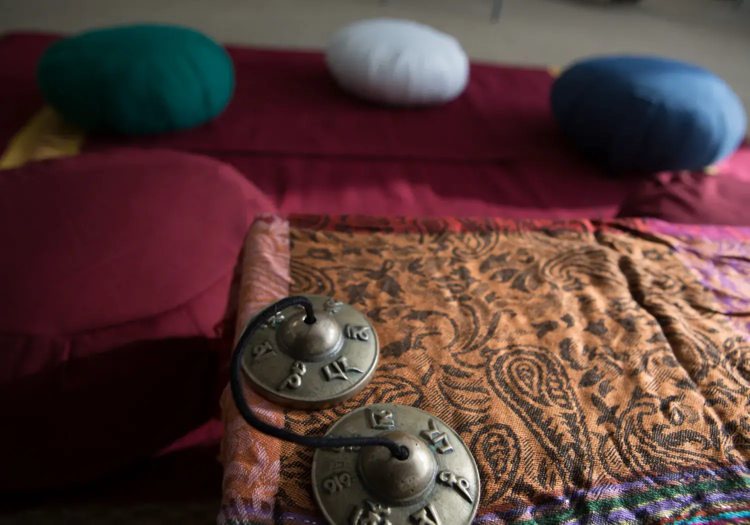 A tranquil meditation space is shown, with a close-up focus on a pair of small, brass cymbals resting on a paisley-patterned fabric. Behind them, soft meditation cushions (zafus) in various colors sit on a red mat, ready for use.