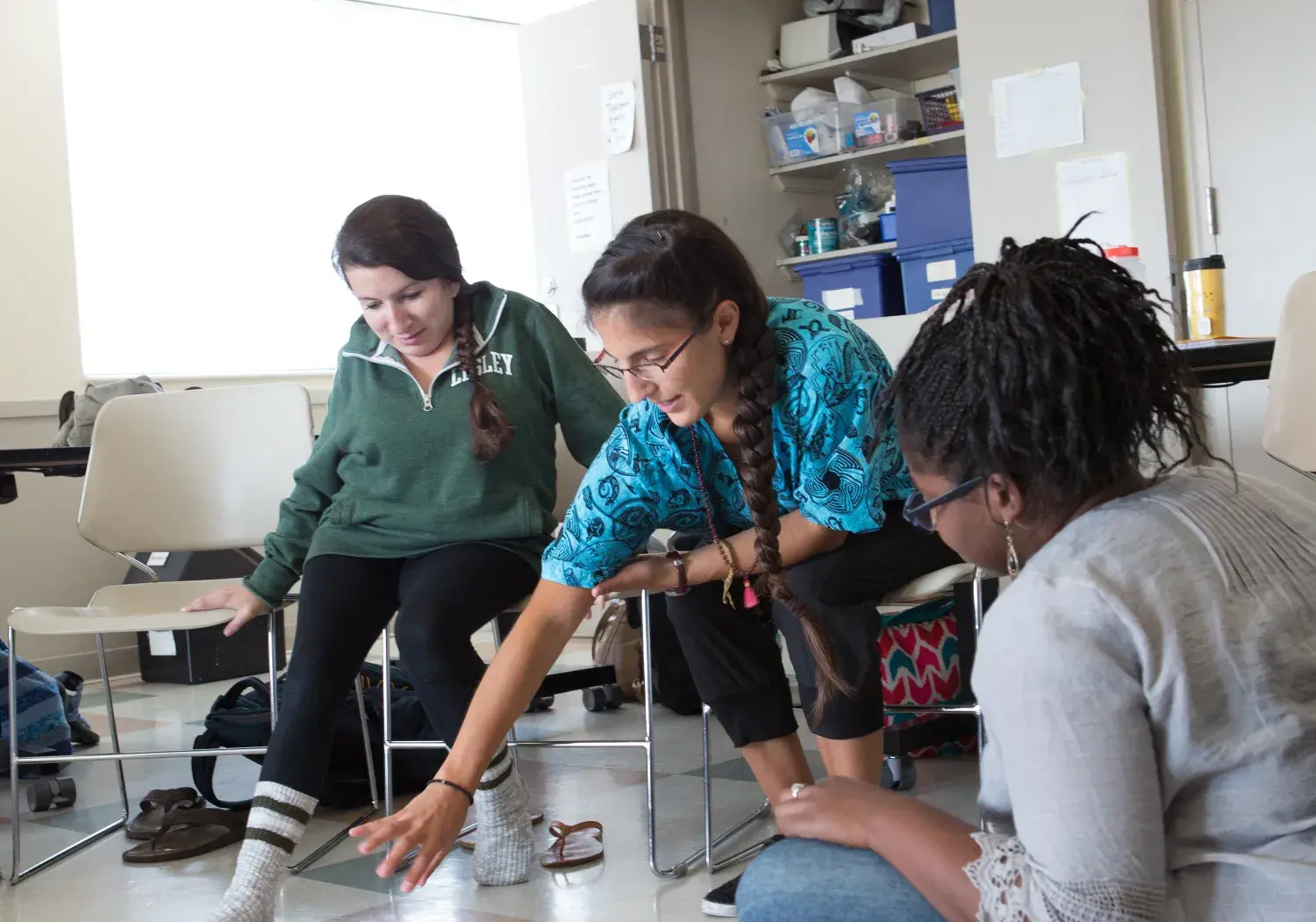Three participants in an art class or workshop have an informal critique. With artwork displayed on the floor between them, one woman points to a drawing, engaging the other two in a conversation about the pieces.