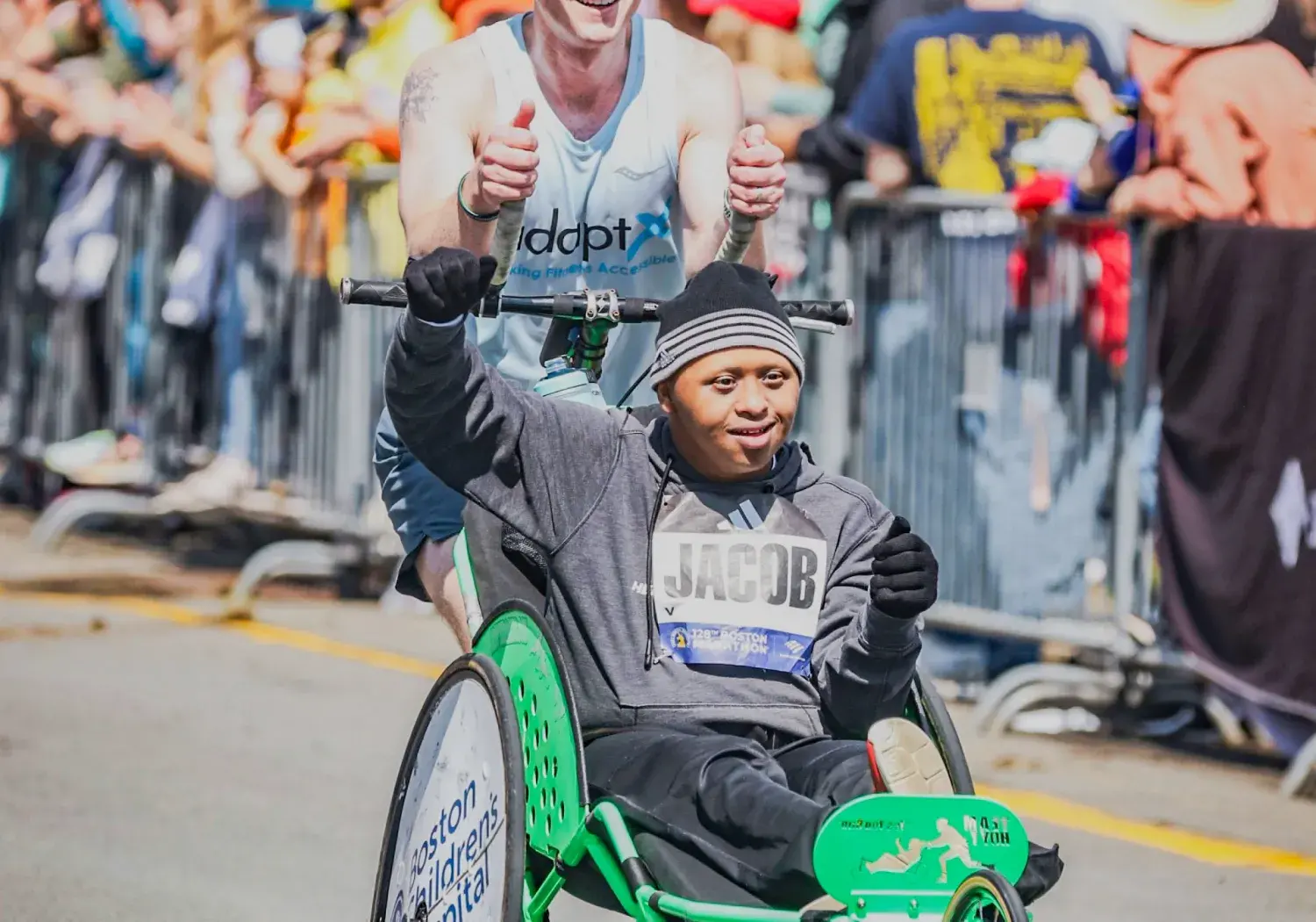 A smiling runner gives two thumbs up while pushing a young athlete named Jacob in a bright green adaptive racing wheelchair during a race. They are surrounded by cheering spectators behind metal barricades on a sunny day. Both individuals radiate joy and determination.