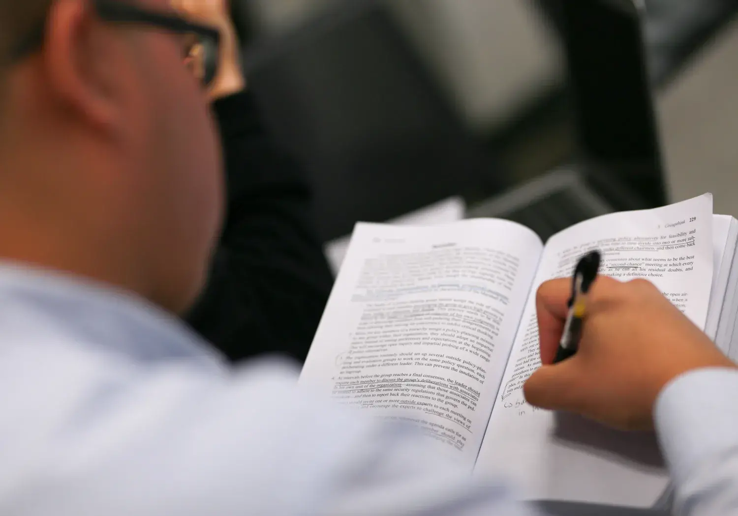 A student with glasses is closely reading a textbook, holding a pen and making handwritten notes in the margins. The page contains printed text with some underlining and annotations. The image is focused on the book and pen, with the student slightly blurred in the foreground. A laptop is partially visible on the desk.