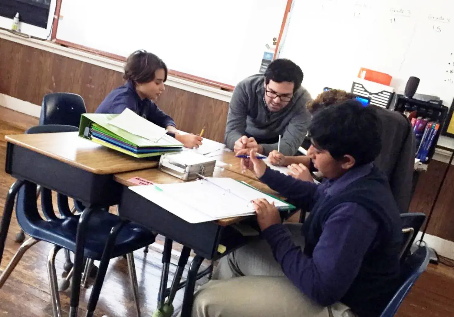 A classroom scene shows three students seated around desks pushed together, engaged in group work or a collaborative assignment. An adult—likely a teacher or facilitator—leans in to assist, interacting closely with the students. The group is focused on paperwork and notebooks on the desk. The room features hardwood floors, a globe on a stand in the background, and a whiteboard with writing on the far wall, indicating an academic environment. The atmosphere suggests positive engagement and active learning.