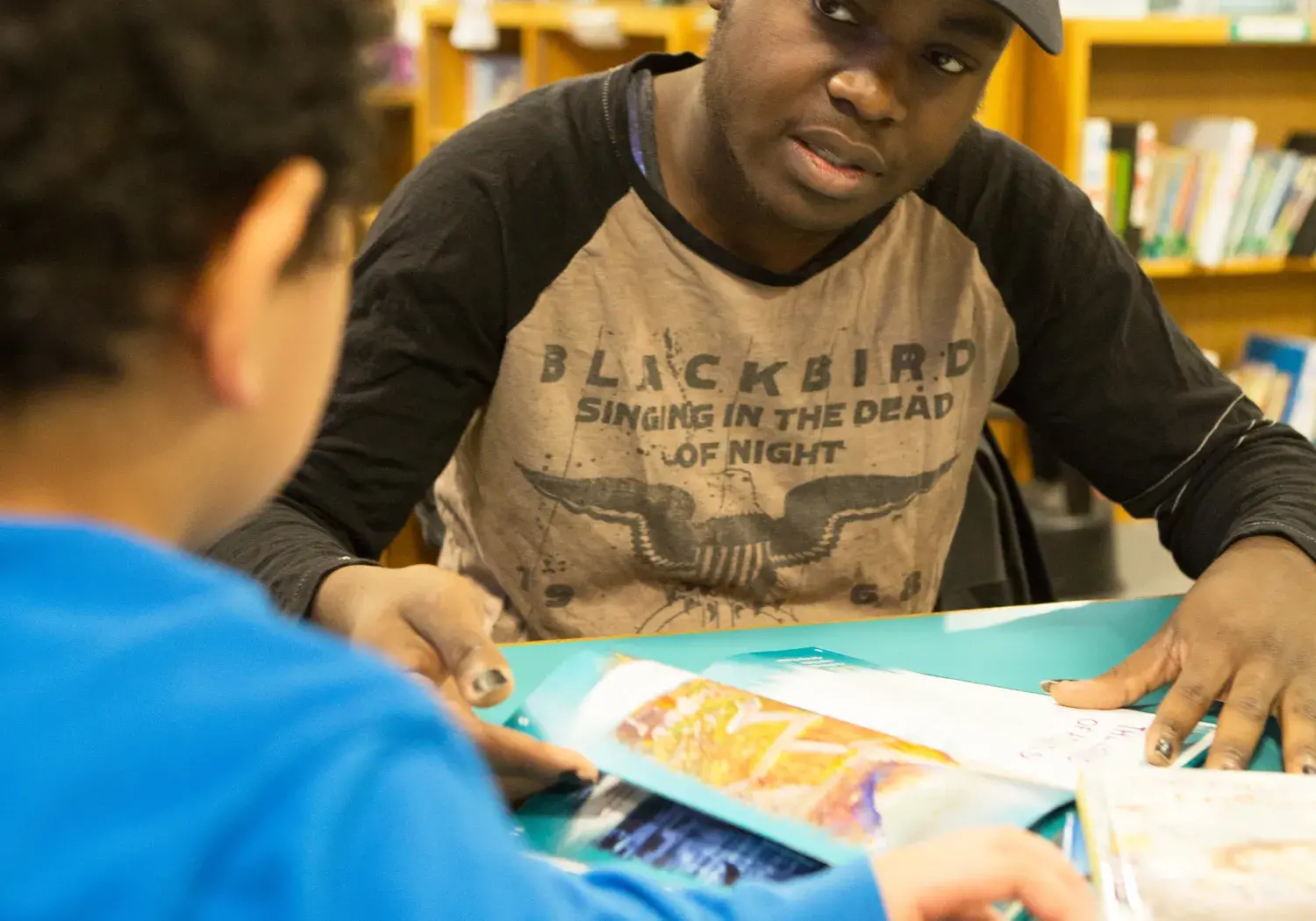 A person wearing a black cap and a shirt with the text "BLACKBIRD SINGING IN THE DEAD OF NIGHT" sits at a table in a library, interacting with a child in a blue shirt. They are looking at colorful, open books spread out on the table, with bookshelves and a globe visible in the background. The setting appears to be an educational or reading activity.