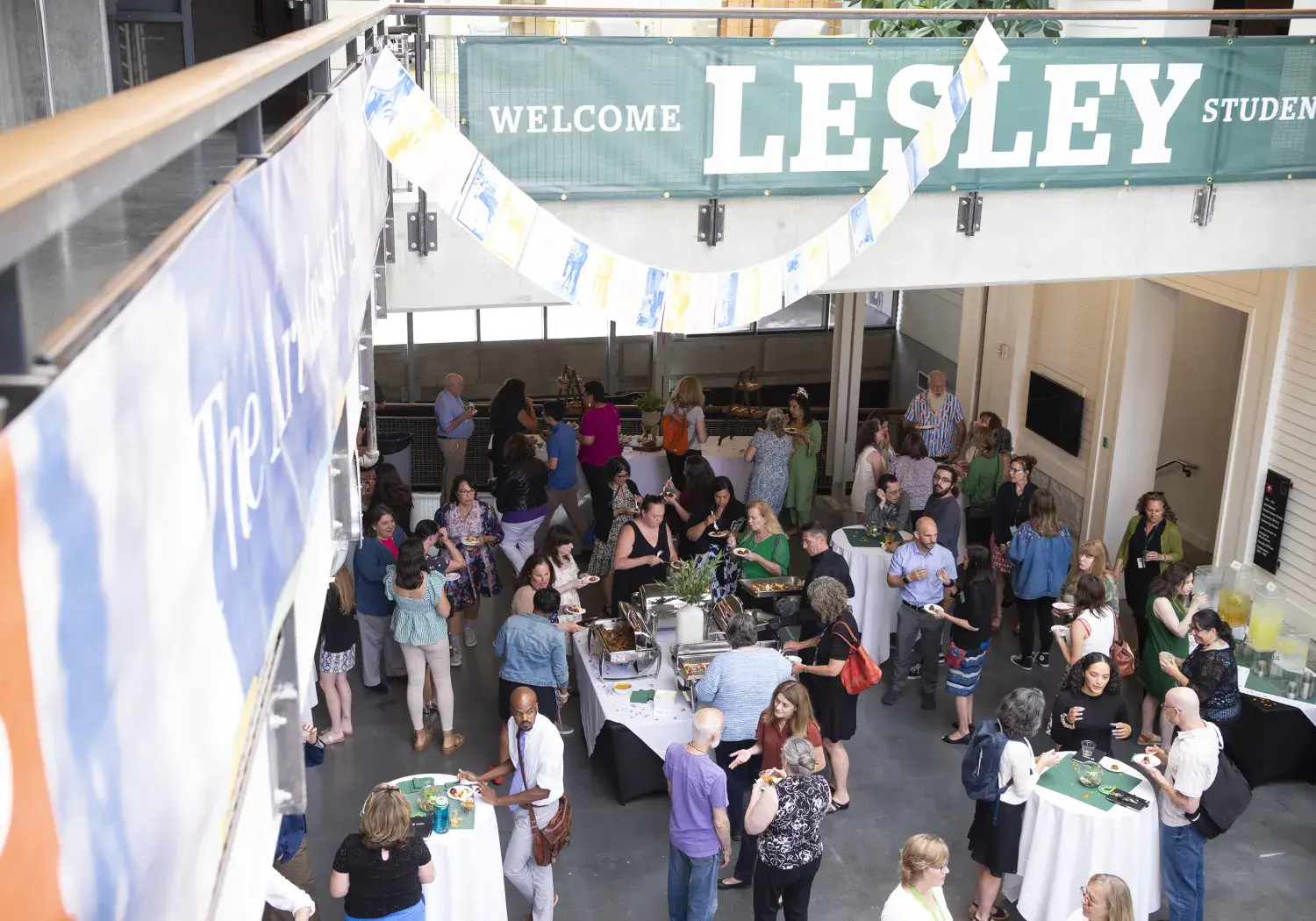 An aerial view of a bustling indoor event with a large group of people gathered around tables. A long green banner with "WELCOME LESLEY STUDENTS" in white letters is strung across the top of the space. Colorful flags are draped below the banner. People are interacting, some are serving themselves from buffet tables, and others are holding drinks. Large banners are hanging from a railing on the left side of the frame.