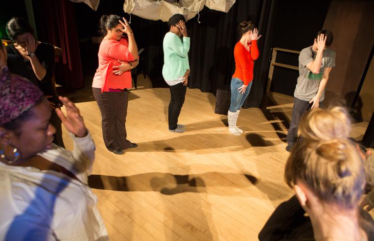 People in a circle on a stage during a drama therapy exercise stand with one hand up against their faces.