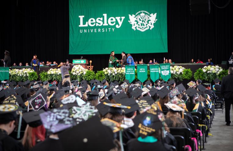 Photo of commencement stage with big green bannister that says Lesley. People sitting with diplomas in chairs facing the stage