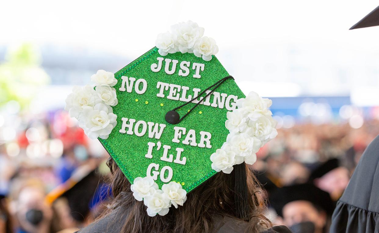 A graduate from the 2022 Commencement with a decorated cap. The cap is green with white flowers on its four corners and says "Just No Telling How Far I'll Go."