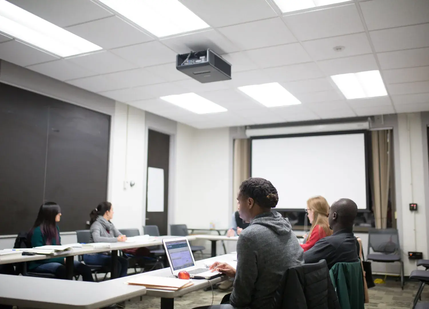 Photo of six students sitting in a square shape at tables in a classroom. Main student in focus is engaged in materials on her laptop.