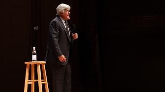 Jay Leno standing on Symphony Hall stage with a stool in the foreground