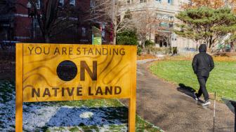 Sign: "You are standing on Native Land" - installed outside, person walking by.