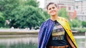 Ashley Delgado with a river and buildings behind her, wearing a cape.
