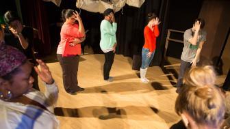 People in a circle on a stage during a drama therapy exercise stand with one hand up against their faces.