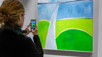 A woman takes a photo of a painting with her cell phone.