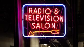 Neon sign that reads: "radio and television station" with an arrow pointing to the bottom left corner of the rectangular sign.