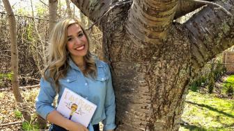 Katelyn Twardzik holding a children's book and leaning against a tree.