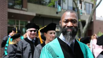 Matthew E. Henry wearing graduation robes with other people in the background. Standing outside.