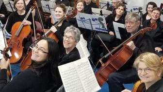 An orchestra seated in with men and women holding instruments including violins, cellos and clarinets.