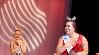 Sarah Achorn holds her hand to her heart in excitement as she wears the crown and Miss New Bedford sash on stage in her floor-length red dress.