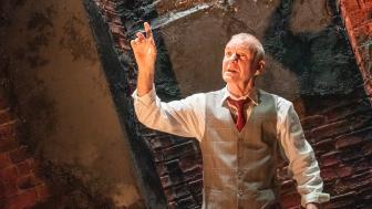 Man in a suit vest and pants holding up one hand, with a brick wall behidn him.