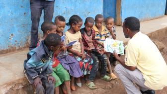 Children in Ethiopia sitting on the sidewalk while their teacher shows them a book.