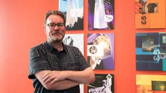 Graphic designer Vaughan Oliver stands with arms crossed and a slight smile in front of posters he's designed.