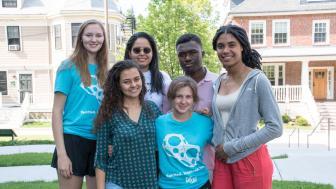 Seven students stand in a group picture on Doble Quad with two Victorian houses behind them.