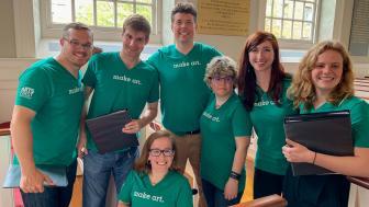 Group of people sitting in or standing around pews wearing green shirts that say "Make Art"