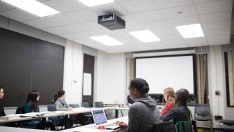 Photo of six students sitting in a square shape at tables in a classroom. Main student in focus is engaged in materials on her laptop.
