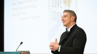 Richard Blanco standing at a podium with hands clasped and smiling.
